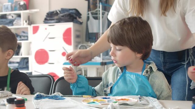 A caring young art teacher assists a boy in creating drawings on clothing during a master class.