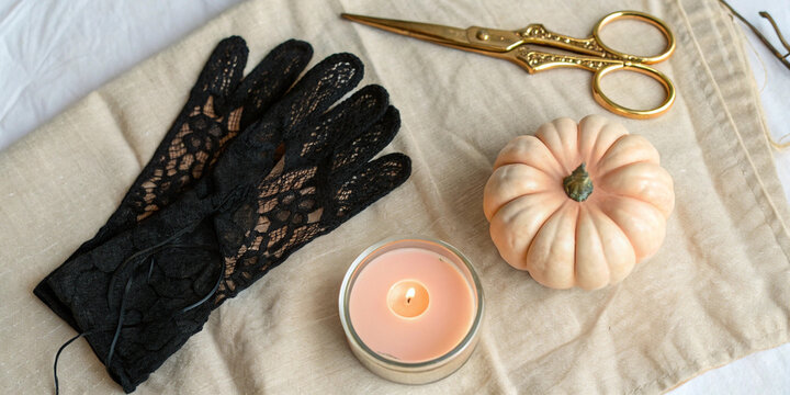 Halloween themed still life featuring black lace gloves, a burning scented candle, a small pumpkin, and golden scissors on a beige linen cloth, creating a cozy atmosphere - Powered by Adobe