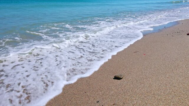 Small pebble on sandy beach touched by gentle turquoise waves of Aegean Sea under bright summer sun. Kos. Greece.