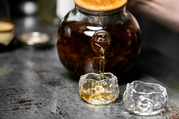 Close-up of tea being poured from a glass teapot into a small transparent cup on a rustic surface, highlighting warm amber tones and elegant glassware details.