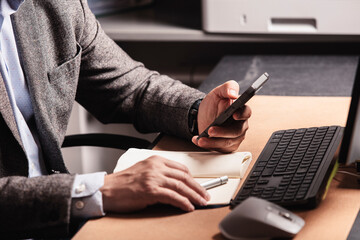 Businessman working at modern office desk, holding smartphone and pen, writing in notebook, with...