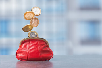 Coins drop from above into a red purse against a blurred background of a city setting during daytime