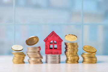 Model house stands among stacks of coins in an indoor setting during daylight, symbolizing investment in real estate and finances