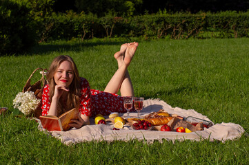 young blonde woman in a red floral dress having a picnic in the park, lying on a plaid with a...