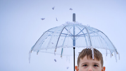 A young boy stands outdoors holding a transparent umbrella covered in ice, as snow drifts down softly around him.