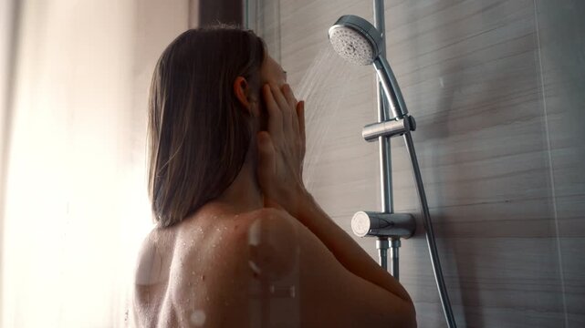 Young woman taking shower in modern glass shower cabin and wash face. Girl washing under running water in hotel or at home.