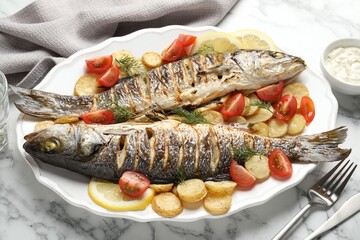 Tasty roasted seabass fish served with potatoes, tomatoes, sauce and lemon on white marble table, closeup. Homemade seafood dish