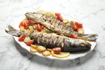 Tasty roasted seabass fish served with potatoes, tomatoes and lemon on white marble table, closeup. Homemade seafood dish
