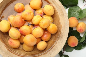 Fresh ripe apricots in bowl with water and green leaves on white table, flat lay