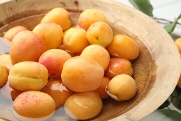 Fresh ripe apricots in wooden bowl with water and green leaves on table, closeup