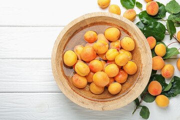 Fresh ripe apricots in bowl with water and green leaves on white wooden table, flat lay. Space for text