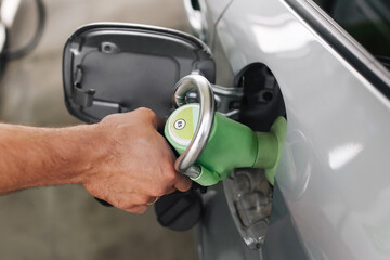 Fueling a car with green nozzle. Close up of man&rsquo;s hand holding fuel dispenser. Refueling vehicle at gas station. Eco friendly transport concept. Clean silver car with open fuel tank flap.