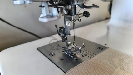 A close-up of a sewing machine, focusing on the needle and presser foot. The machine is white with a metallic needle plate, ready to stitch fabric.
