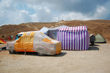 Improvised colorful camping setup with car covered in tarp at dry summer hillside campsite. Camping, festivals, alternative travel, sustainable tourism, outdoor life, survival, vanlife, boho adventure