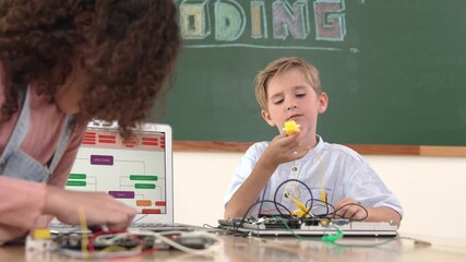 Caucasian energetic boy fixing main board and looking at camera while american student working together to inspect electronic machine by using color wires at table with laptop display code. Pedagogy. - Powered by Adobe