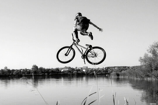 Black and white photo of a person performing a high jump with a bicycle over a river. The boy is captured mid-air in an acrobatic pose against a clear sky and calm water. Freedom, adventure, youth