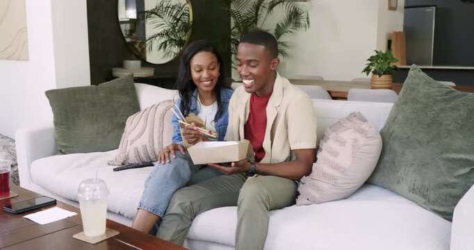 African American couple settling on sofa at home eating takeout using chopsticks laughing at screen
