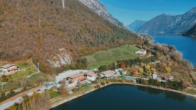 Drone shot of Lake Idro with hillside homes, autumn trees, green meadows, and blue water