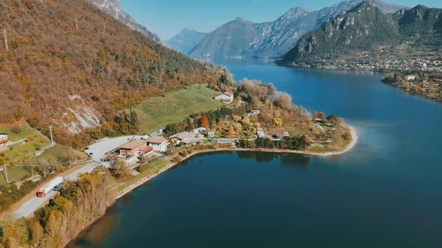 Drone shot of Lake Idro with hillside homes, autumn trees, green meadows, and blue water