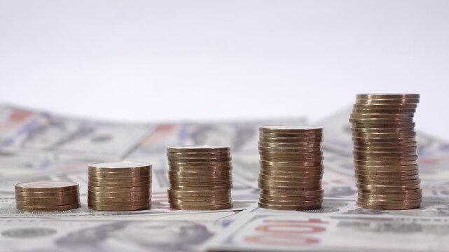 Four stacks of coins increasing in height with a hand placing a coin on top, set on a backdrop of dollar bills, evoking wealth and success.
