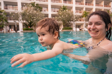 Caucasian woman holds her son while swimming in a pool on vacation. Mom teaches little boy to swim. 