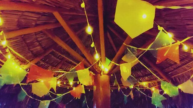 Colorful flags and warm string lights decorate a rustic straw hut roof for a traditional brazilian June Festival. Festive night celebration ambiance with typical party decorations hanging.