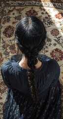 A girl with a dark braided hairstyle sits on an ornate floral carpet, wearing a navy dress speckled with white, viewed from above in soft daylight.