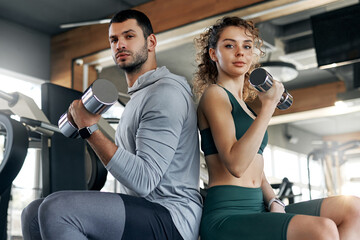 Confident man and woman sitting back to back while holding dumbbells in modern gym. Strength, unity, and focus on fitness goals together.