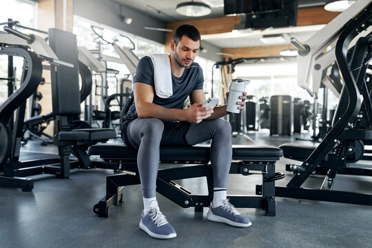 Young athletic man sitting on bench in gym, checking smartphone and holding protein shaker after training session. Concept of post-workout rest and fitness routine.