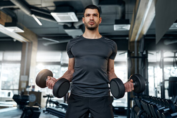 Young athletic man performing biceps curls with dumbbells in a modern gym. Strength and...