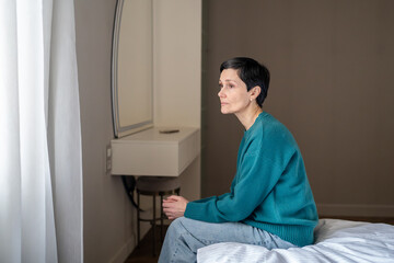 Depressed drained woman sitting on bed staring out window arms crossed. Agoraphobia, social fear anxiety, isolation, emotional exhaustion, waiting nervously, mental health struggle, indoor confinement