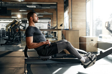 Focused athletic man using a rowing machine during workout at a bright, spacious gym. Concept:...