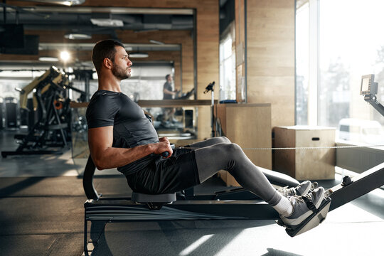 Focused athletic man using a rowing machine during workout at a bright, spacious gym. Concept: endurance, cardio, fitness, health, discipline.