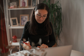 Focused woman in glasses writing notes at home workspace with laptop, seated at desk in cozy room...