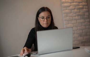 Serious woman in glasses working on laptop at minimalist desk, using mouse, focused on screen, with...