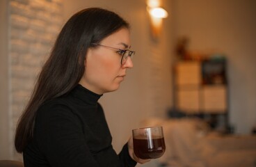 Side view of woman in glasses holding glass cup of tea or coffee, sitting calmly in cozy home...