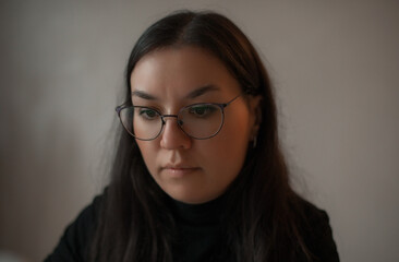 Close-up portrait of a thoughtful woman in glasses with long dark hair, looking down with focus in...