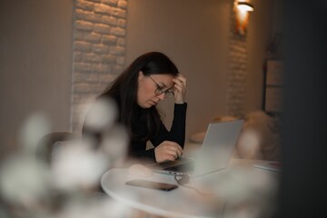 Woman in glasses sitting at home office desk, looking stressed while working on laptop, hand on...