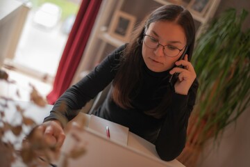 Young woman in glasses multitasking at home, talking on phone and using laptop at desk with...