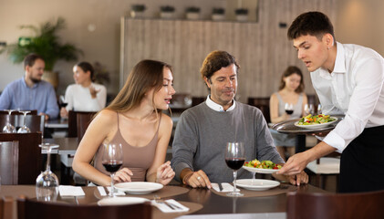 Male waiter serving fresh vegetable salads to married couple guests visiting restaurant for dinner, sitting at table with glasses of red wine