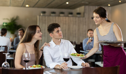 Friendly waitress bringing dishes salad to guests of cozy restaurant