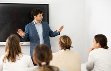 Positive male trainer, conducting advanced training courses to office employees sitting at desks in...