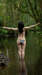 Fototapeta premium Woman on her back in a wooded stream, with arms raised in a pose of yoga or meditation. Submerged up to the thighs, it reflects peace and well-being. Dense green foliage creates a natural environment,