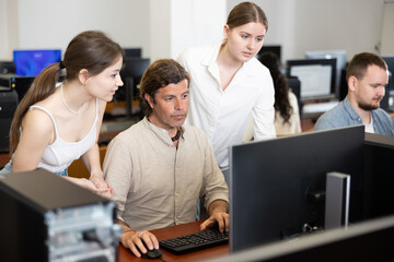 Two female friend companions stands next to computer courses male student, looks at monitor screen...