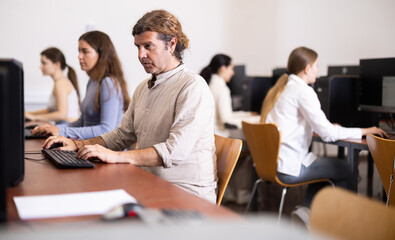 Man student of computer courses attentively looks at monitor screen, types on keyboard and...