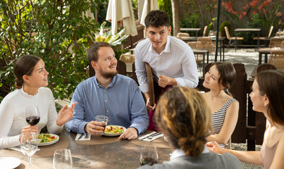 Young male waiter takes order from group of male and female friends on restaurant terrace