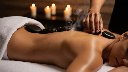 Medium shot of a therapist placing hot steaming basalt stones on a woman's back for a relaxing massage in a spa with soft candlelight. 