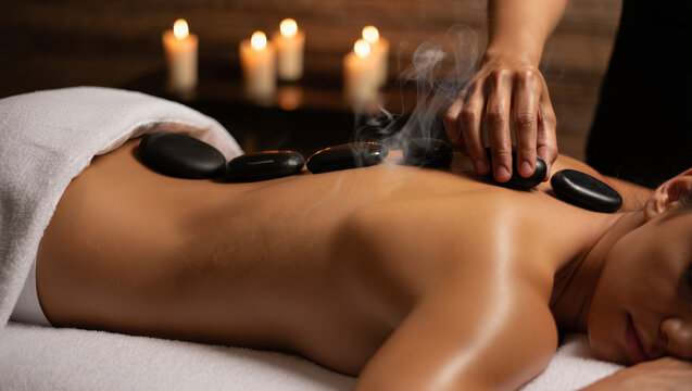 Medium shot of a therapist placing hot steaming basalt stones on a woman's back for a relaxing massage in a spa with soft candlelight.
