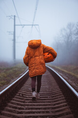 Girl in orange jacket without head with carved pumpkin jack-o-lantern walking on foggy railway...