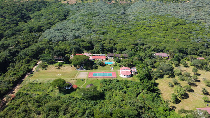 Aerial view of a resort with a pool and tennis court surrounded by lush green trees and vegetation...